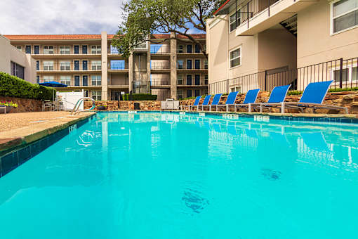 A pool area with lounge chairs, a patio table and chairs with a blue umbrella and stainless steel BBQ in front of apartment buildings at Princeton Court Apartments in the Vickery Midtown neighborhood of Dallas, TX.