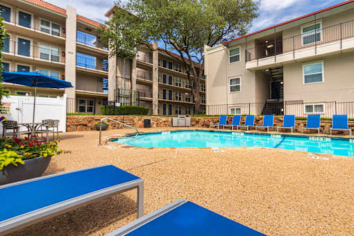A pool area with lounge chairs, a patio table and chairs with a blue umbrella and stainless steel BBQ in front of apartment buildings at Princeton Court Apartments in the Vickery Midtown neighborhood of Dallas, TX.
