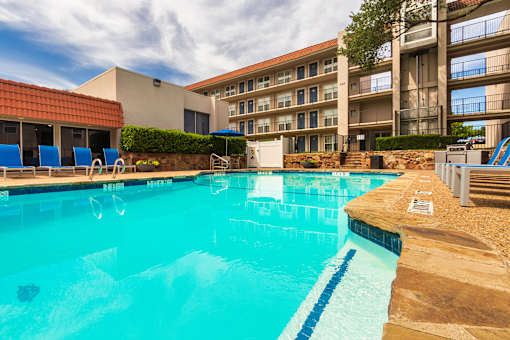 A pool area with lounge chairs, a patio table and chairs with a blue umbrella and stainless steel BBQ in front of apartment buildings at Princeton Court Apartments in the Vickery Midtown neighborhood of Dallas, TX.