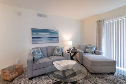 A modern living room with a grey sofa, chaise lounge and a glass coffee table in the 558 square foot 1 bedroom, 1 bath model apartment at The Summit at Midtown Apartments in the Dallas Midtown neighborhood of Dallas, TX.