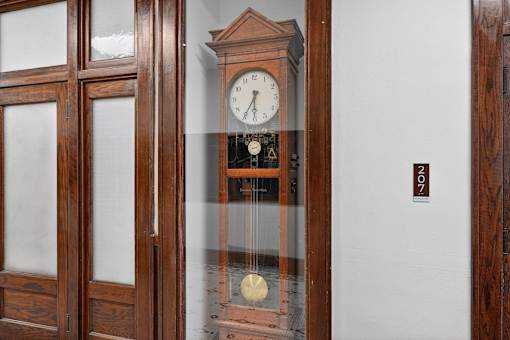 A wooden clock with a glass case is on a white wall.
