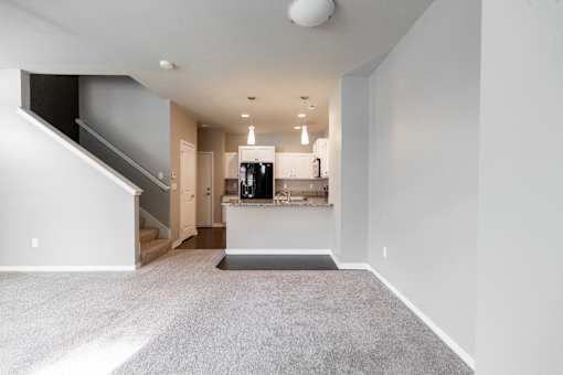 A spacious, empty room with a staircase on the left and a kitchen area in the background.