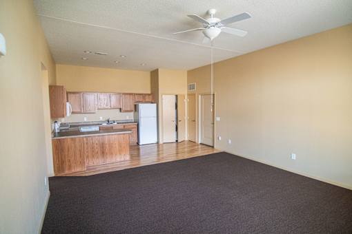 Empty room with a ceiling fan and a kitchen area in the background.at Cottages at Johnston Commons, Johnston, IA  