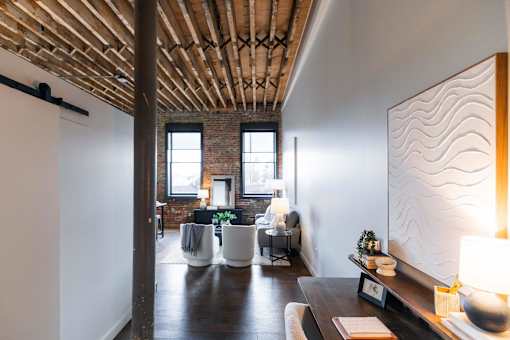 A room with a wooden ceiling and a white wall with a lamp on a table.at Plant 1 Lofts, N. Newton  
