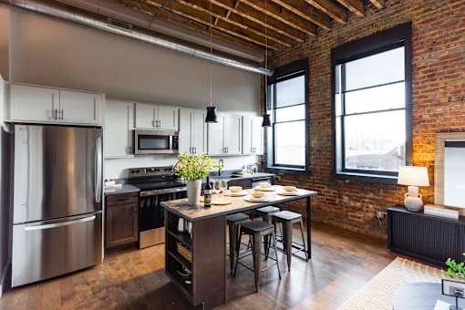 A kitchen with a wooden table and chairs, a refrigerator, and a brick wall.at Plant 1 Lofts, N. Newton  
