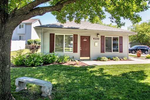 A small house with a brown door and windows is surrounded by greenery at Hunters Run Apartments, Newton, Iowa