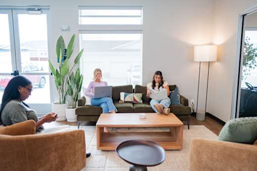 Three women are sitting in a waiting area with a laptop and a book.