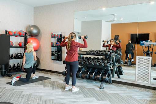 A woman in a red top and black leggings is working out with dumbbells in a gym.