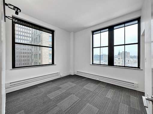 A room with a grey carpet and two windows overlooking a cityscape at The Des Moines Building Apartments, Des Moines, Iowa