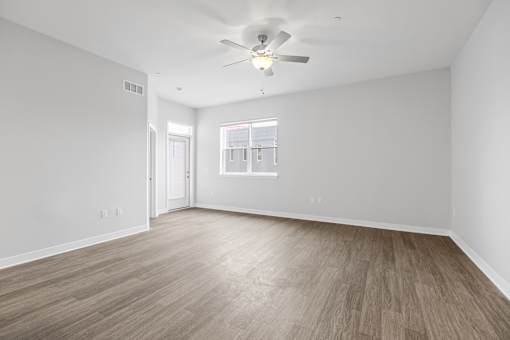 A room with a ceiling fan and light, wooden flooring, and a window with a view of a building outside.