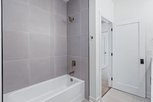 Modern bathroom with white tub and gray tile surround at The Flats at West Glen Apartments, West Des Moines, IA