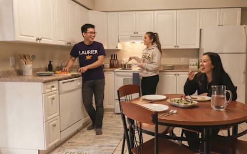 A man in a purple shirt is standing in a kitchen with a woman in a grey hoodie and another woman sitting at a table.