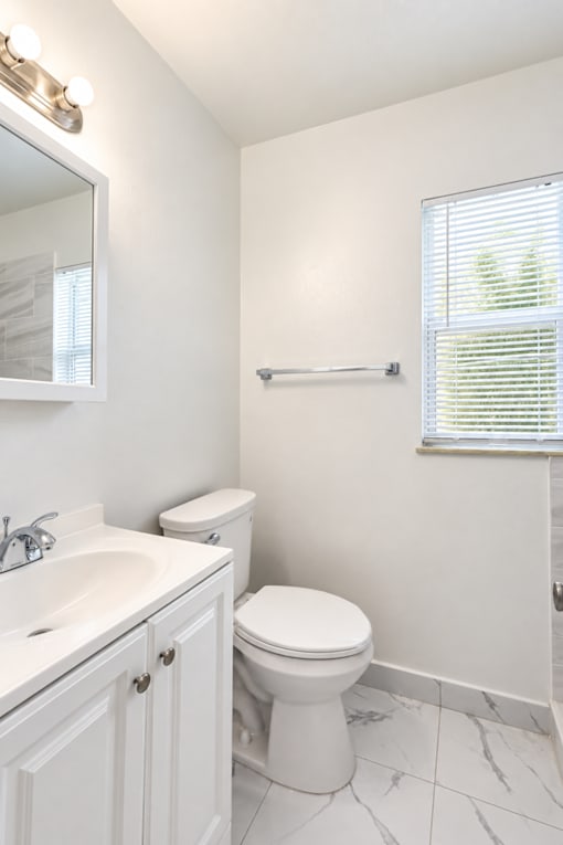 A white bathroom with a toilet, sink, and a window with blinds.