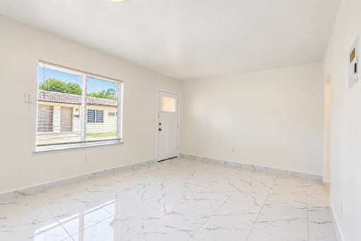 A room with white walls and marble flooring with a window showing a view of a house.