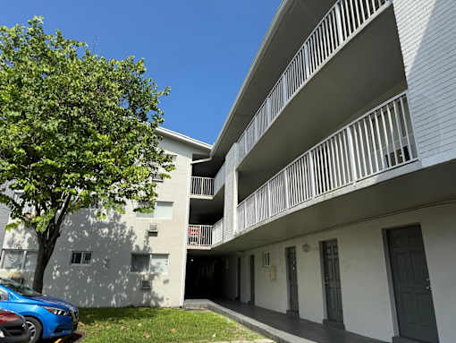 A blue car is parked in front of a white building with a tree to the left.