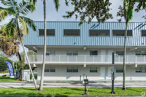 A white building with a blue stripe on the side is surrounded by palm trees.