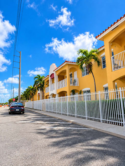 A car is parked on the side of a street in front of a yellow building with a white fence.