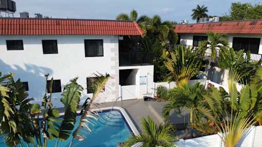 A white building with a red roof and a pool surrounded by palm trees.