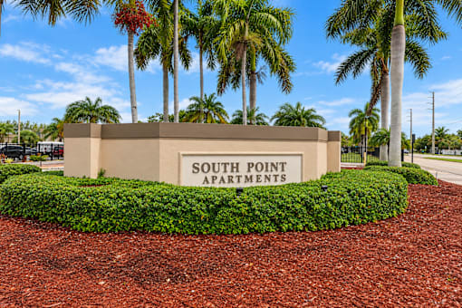 A sign for South Point Apartments is surrounded by a hedge and palm trees.