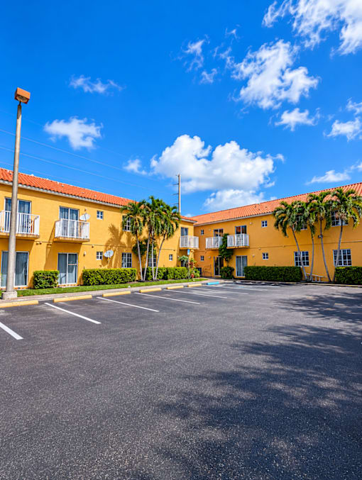 A sunny day at the parking lot in front of a yellow building with palm trees.