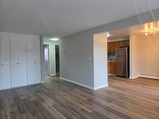 A room with wood flooring and a kitchen in the background.