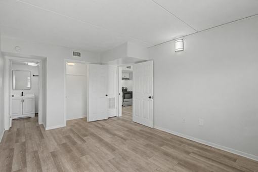 A white room with wood flooring and a kitchen area in the background.