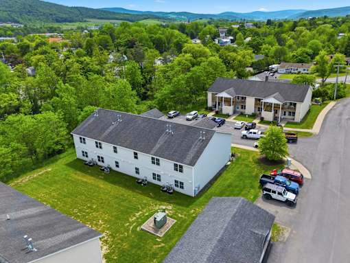 A white house with a black roof is surrounded by greenery and a parking lot.