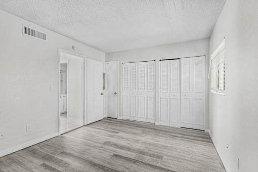 A room with white walls and wood flooring with a door on the left and three closets on the right.