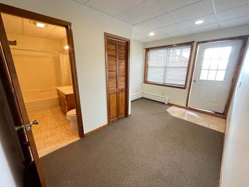 A room with a carpeted floor, a toilet visible through the open door, and a window with blinds.