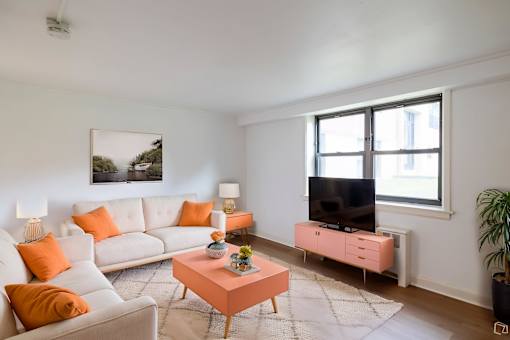 A living room with a white couch, orange pillows, a pink coffee table, and a flat screen TV.