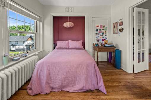 A bedroom with a pink bedspread and a window with a view of houses.