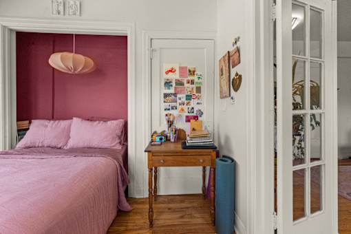 A bedroom with a pink bed and a table with a lamp on it.
