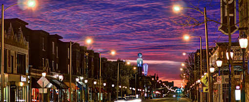 A city street at dusk with buildings on either side and street lights on.