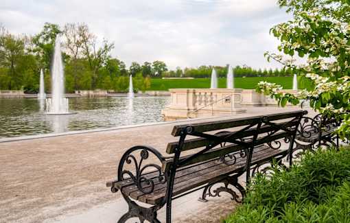 A bench with a metal frame is in the foreground of a park with a fountain and trees in the background.