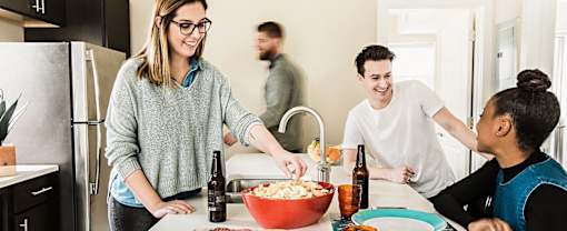 A woman in a grey sweater is serving popcorn to a man and a woman in a kitchen.