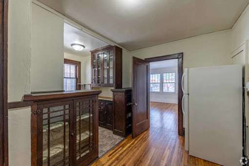 A white refrigerator is in the corner of a room with wood floors and white walls.