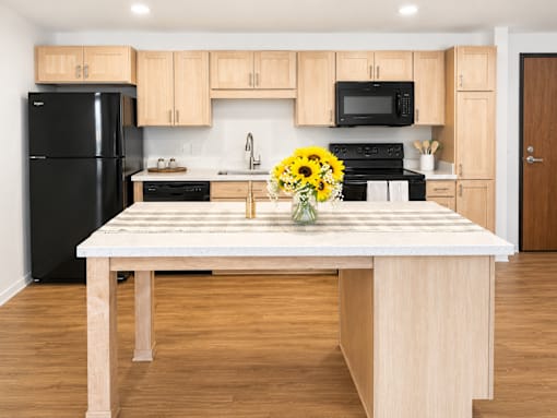 A kitchen with a black refrigerator, wooden cabinets, and a wooden island with a vase of yellow flowers on it.