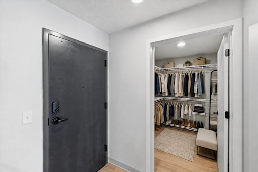 A black door is on the left of a room with a white wall and a white closet with clothes hanging on it.