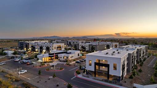 A modern building complex is lit up at dusk with cars parked in the lot.