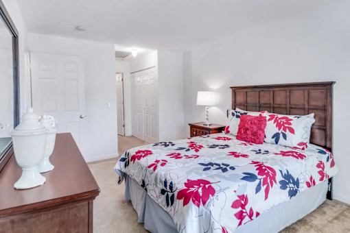 A bed with a floral print is in the center of a bedroom with a wooden dresser and a white vase on top.