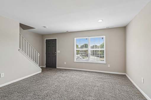 A spacious room with a carpeted floor, a staircase, and a large window overlooking a parking lot.