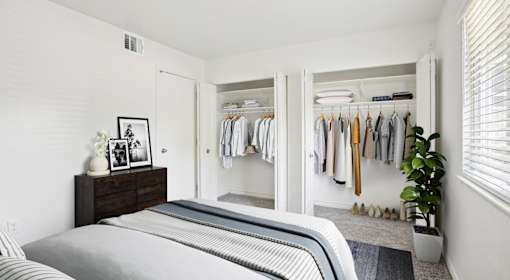 A bedroom with a bed, a dresser with a picture frame, a closet with clothes, and a potted plant.