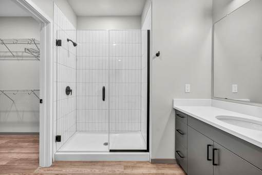 A white tiled shower with a walk-in shower door is in a bathroom with a white sink and a white vanity.