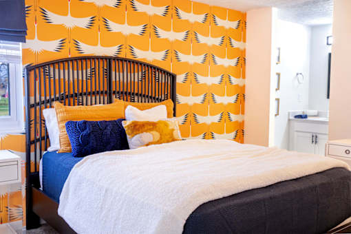 A bed with a white comforter and a blue pillow is in front of a wallpapered wall.