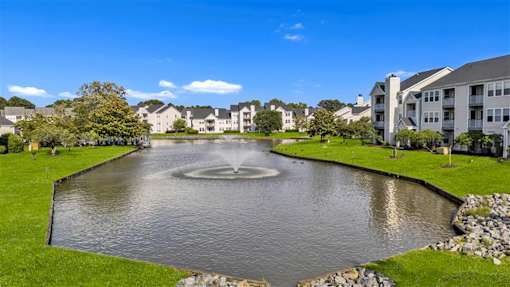 A fountain in the middle of a pond surrounded by grass and buildings.