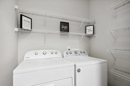 Two white washing machines in a laundry room with shelves above them.