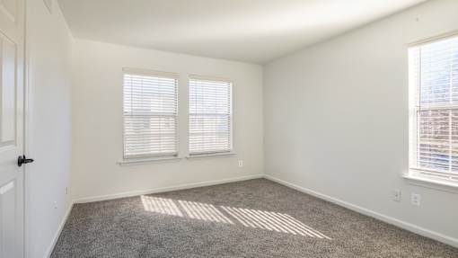 A room with a carpeted floor and two windows with blinds.