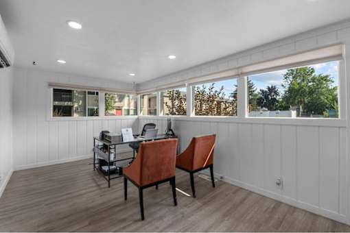 A room with a desk and chairs with a window in the background.