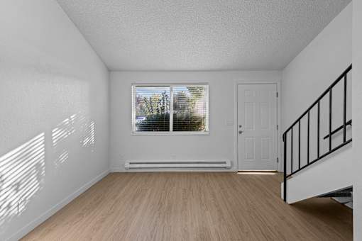 A room with a wooden floor, a window with blinds, a white door, and a staircase with a black railing.