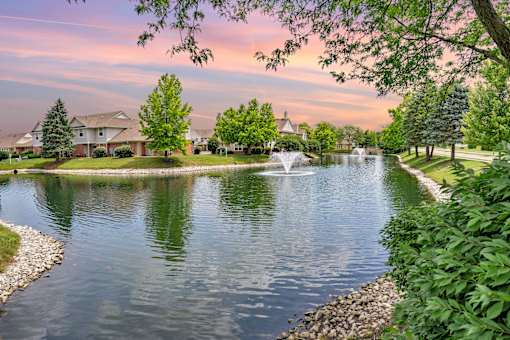 A serene pond with a fountain in the middle surrounded by lush greenery and residential houses in the background.